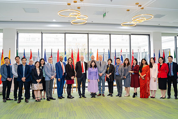 a group of 21 men and women standing in front of many flags from different nations and large windows with a large building behind them.
