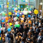 a crowd of people celebrating in an atrium with balloons of all colors falling from the ceiling.