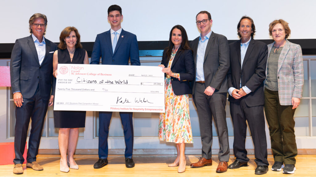 Lucas Suero stands on stage with an oversized check next to judges, PIHE codirectors, and the dean of the Nolan School.