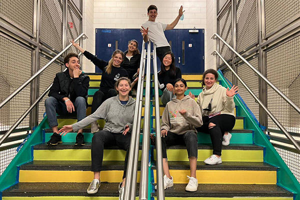 8 young adults sitting on a wide, colorfully painted staircase, smiling and gesturing with their arms at their work.