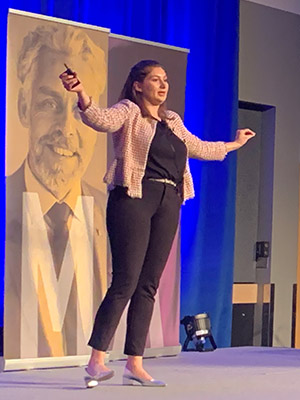 Danielle Boris standing and speaking on a stage with arms open wide open in an inclusive gesture.