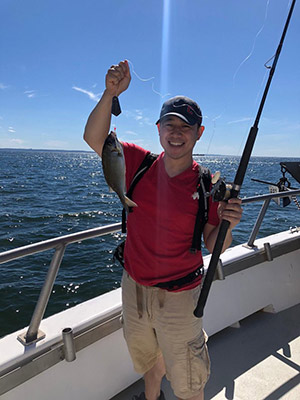 Samuel Cho holding up a fish in one hand and a fishing rod in the other hand with a boat railing and blue ocean and sky in the background.