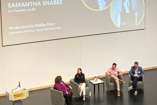 Picture of the moderator and 3 speakers sitting in chairs on the stage with a large screen above them with their names.