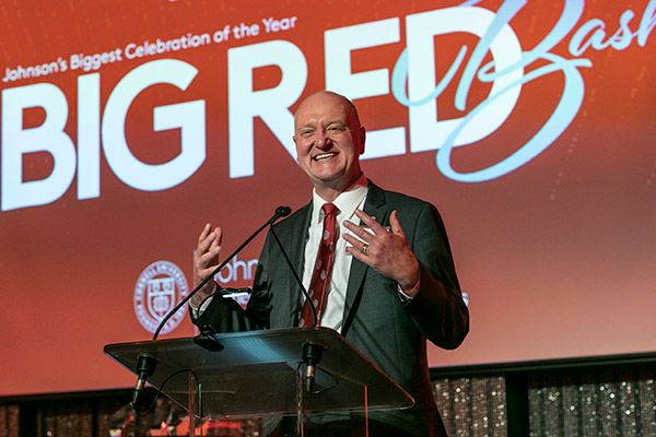 Dean Andrew Karolyi speaking at a podium on stage with the words Big Red Bash on a screen behind him.