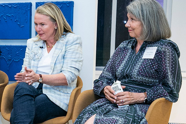 Two women sitting side by side: One is talking intently, looking at the audience and gesturing with both hands, the other is looking on.