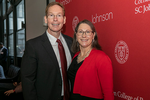 A man and a woman in business attire smiling and standing in front of a red backdrop with the Cornell University and Johnson School logos.