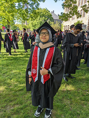 A woman wearing hijab and graduation cap and gown on the Cornell Arts Quad.