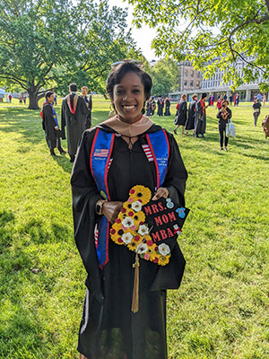 a woman in a graduation gown holding her graduation cap decorated with the words “MBA Mom” and flowers, standing on the Cornell Arts Quad. 
