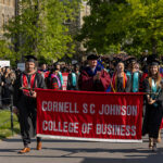 Students and faculty in caps and gowns holding and walking behind a Cornell SC Johnson College of Business banner.