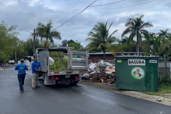 Two men load up a high-capacity truck with branches and tree bark. The truck is parked to the left on the right side of the road and there is a pile of bio-mass debris on the right side of the truck near a dumpster that reads “Basura”.