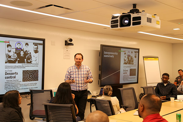 a man stands next to a screen and speaks to students seated at a conference table.