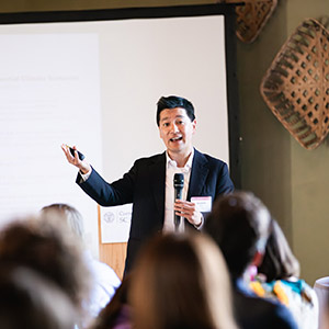 a man standing at the front of a rooming and gesturing as he speaks to the audience.