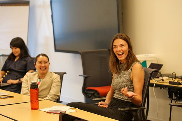 a woman sitting at a conference table with other speaks, smiles, and gestures. 