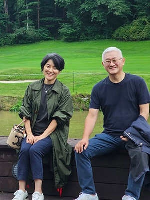 A Korean woman and man dressed in casual clothes sitting side by side on a low wall with a stream, green field, and large evergreens behind them.