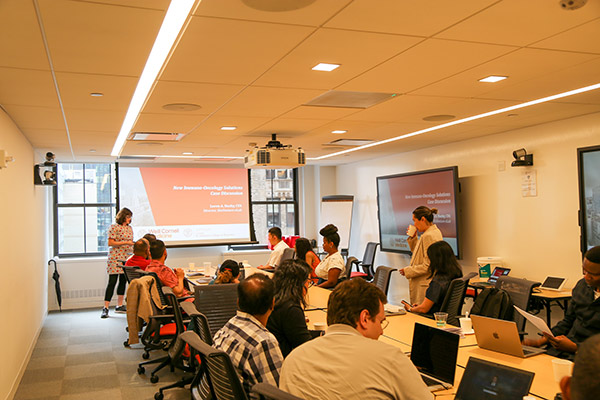 students at a long conference table listening to a woman speaker standing net to a screen at the front of the room.