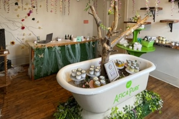 An image of a tub with a tree coming out of it with containers of skin care products in it in the middle of a room with wooden shelves with more products displayed.