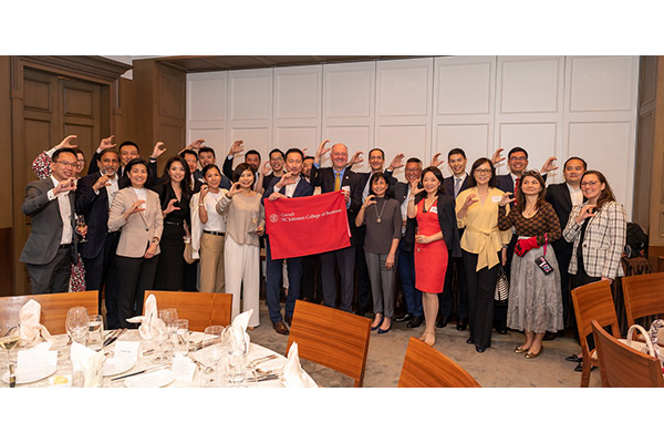 a group of 25 people posing for a photo at the front of a dining room, smiling, and each making a "C" with one hand,