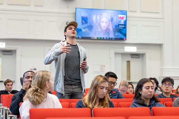 A male student standing and speaking surrounded by other students in a lecture hall.