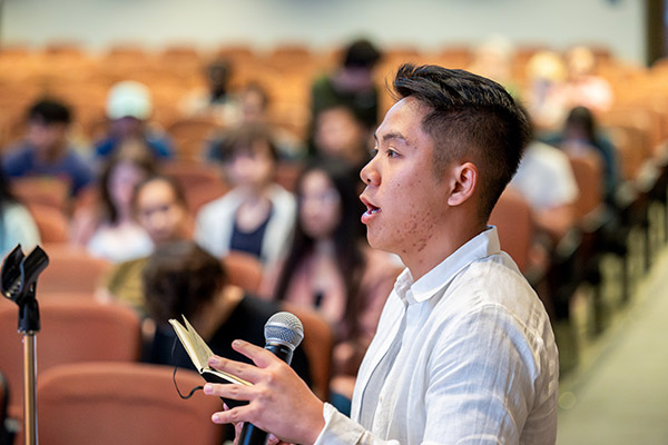 a young man speaking into a microphone with student seated in the background.