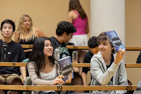 A young man and woman seated in a lecture hall, with other students seated behind them, holding Maxine Bédat’s book, Unraveled.