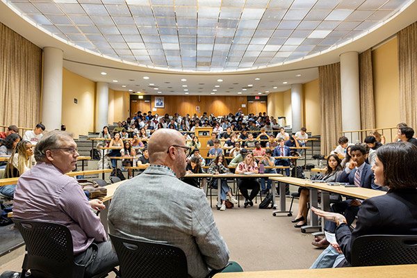 Photo of a large lecture hall filled with students, taken from the front of the room and behind the two faculty members and guest speaker, capturing side views of them.