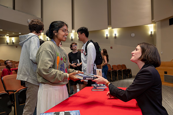 Maxine Bédat seated at a table with student lined up to have her sign their copies of her book. Maxine Bédat hands her book back to a young woman student after signing it.