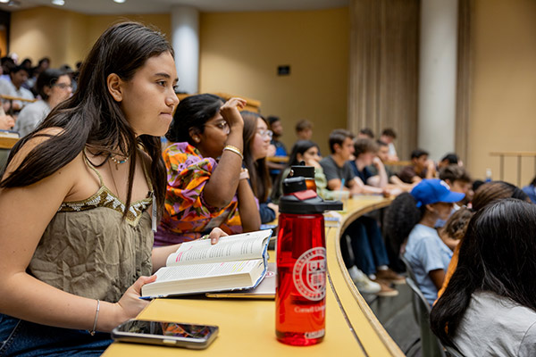 rows of students in a lecture hall, looking engaged; one is raising her hand.