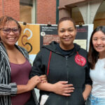 Molebi, JewleKrystal and Sheryl stand in front of a BaldGoddess trifold in Sage Hall's atrium.