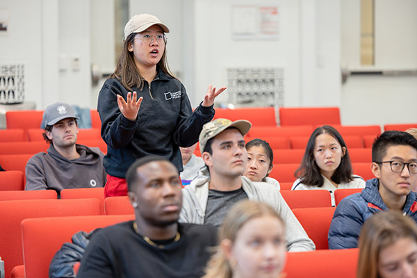 A female student standing and speaking surrounded by other students in a lecture hall.