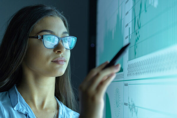 A woman with dark hair and glasses points at a chart on a computer screen with a pen.