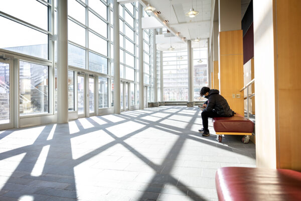 A student sits on a padded bench facing a wall of windows with sunlight streaming inside.