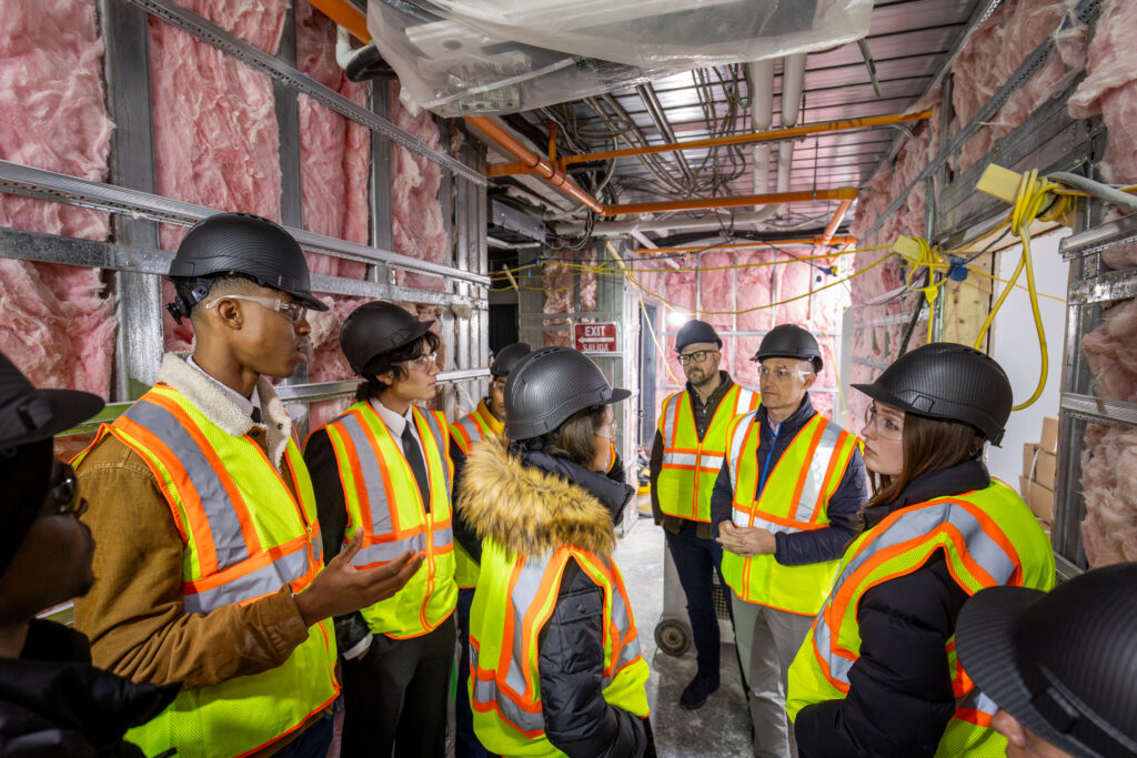 Students wearing hard hats and reflective construction vests gather in an unfinished hallway with exposed insulation.
