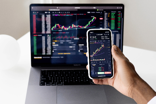Image of a human hand holding a cell phone in front of a laptop with both displaying stock market activities