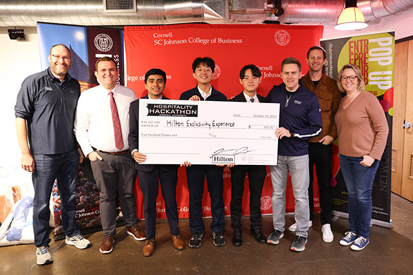 8 people standing on a stage with 4 students holding a novelty check made out to Hilton Exclusivity Experience.