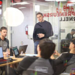 4 students at a table with laptops in a breakout room, one standing, three sitting.