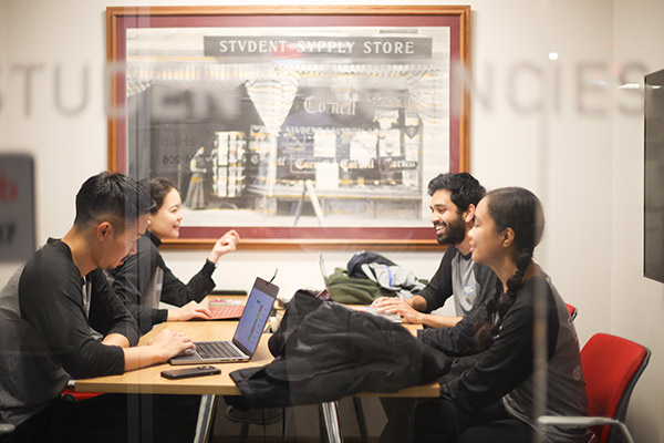 Two male and two female students seated at a table in a glass-walled breakout room with laptops open in front of them. 