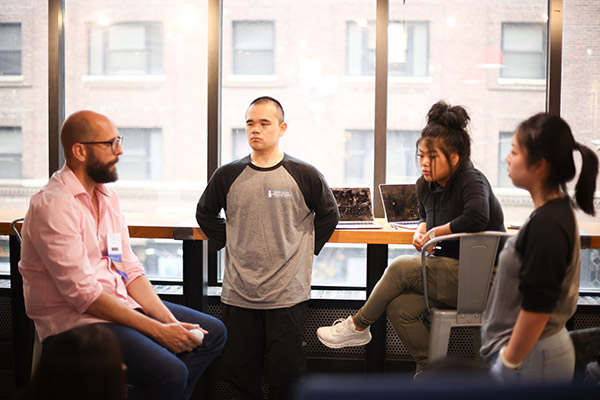 A slightly older man consulting with one male and two female students with a table holding laptops and a window in the background.