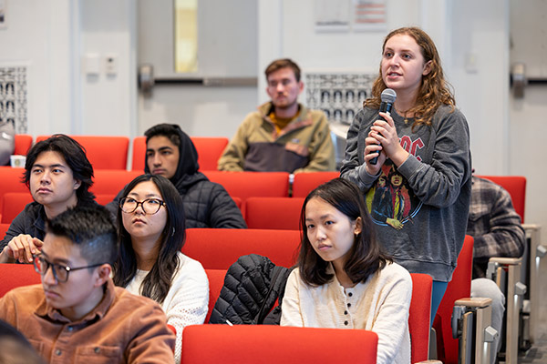 A female student standing at her seat in a lecture hall, surround by seated students, speaks into a microphone.
