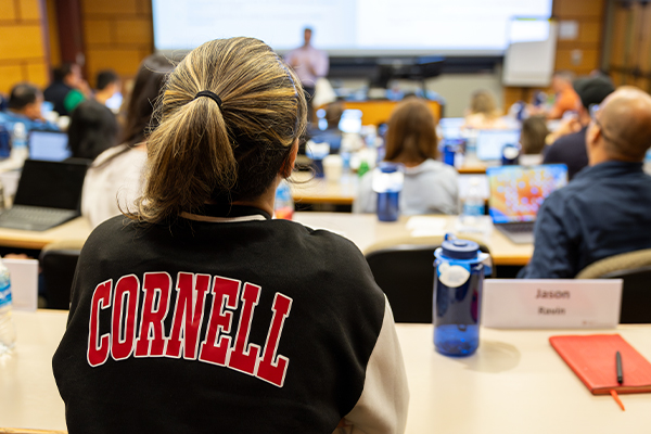 A student wearing a Cornell letterman jacket sitting at the back of a tiered classroom with a professor standing at the front.