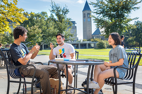Three students sit at an outdoor table with the McGraw Tower in the background.