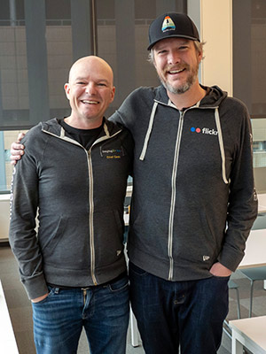 Two mean wearing zip-up hoodies stand smiling with arms around each others' shoulders.