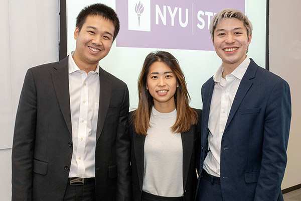 Two male and one female student pose for a photo with the NYU Stern logo on a banner in the background.