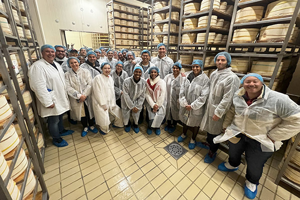 people dressed in white plastic smocks and caps in a room lined with shelves of large cheese rounds.