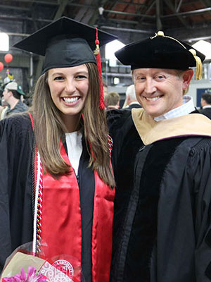 a young woman and an older man standing side by side and wearing graduation regalia / caps and gowns.