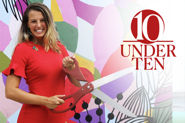 a woman standing in front of a brightly colored background holding a huge pair of scissors cutting a ribbon and smiling. the 10 Under 10 graphic identifier is overlaid on the photo.