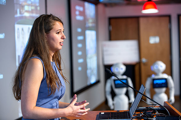 A young woman speaking with a small humanoid robot to her side in the background.