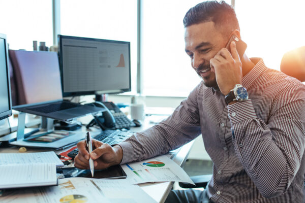 Man on a cell while working at a desk with computers and papers on the desk.