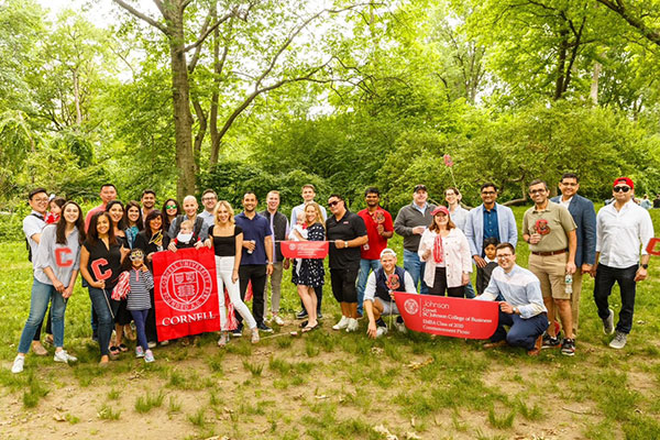 A group of @30 adults, children, and babies standing in a grassy park with trees in the background and holding up Cornell University and SC Johnson College of Business banners.
