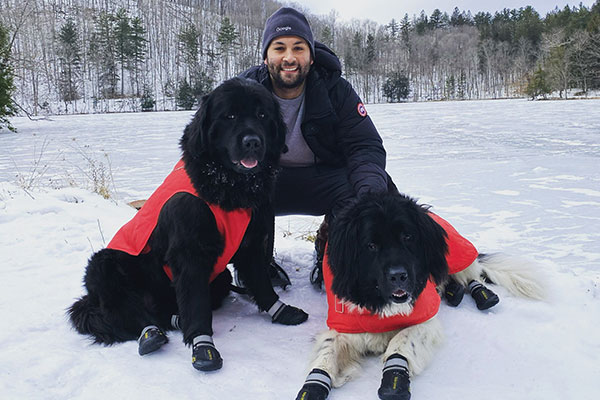 Chris Marino smiling and crouching down in the snow with his hands on two large dogs on either side of him and hills and bare trees in the background.