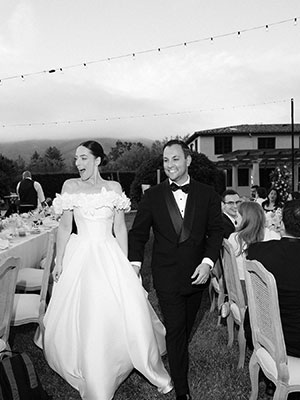A woman in a wedding dress and man in a tuxedo, holding hands and smiling, and walking between tables set up outside, covered with white tablecloths and filled with guests, with trees and hills in the background.